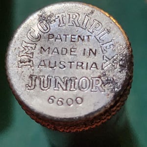 Puede incluir: Primer plano de una tapa metálica plateada con el texto "IMCO TRIPLEX PATENT MADE IN AUSTRIA JUNIOR 6600" en relieve. La tapa tiene un borde texturizado y parece ser vintage.