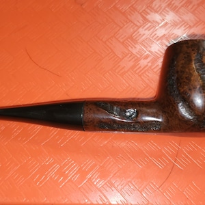 May include: A brown tobacco pipe with a black stem. The pipe has a rounded bowl with carved details and a small, decorative element. The pipe is set against an orange background.