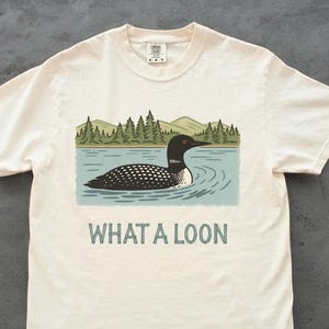 May include: Cream-coloured t-shirt with a graphic of a loon on a lake, with trees and mountains in the background. The text "WHAT A LOON" is printed below the graphic in green. The shirt is made of a soft material.