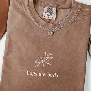 May include: A brown Comfort Colors t-shirt with a white insect graphic and the text "bugs are buds." A silver necklace with a pendant rests on the shirt. The shirt is folded and has a tag.