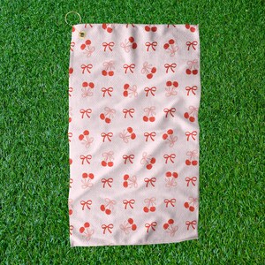 May include: A light pink golf towel with a repeating pattern of red cherries and bows. The towel has a small loop for hanging and is set against a green grass background.