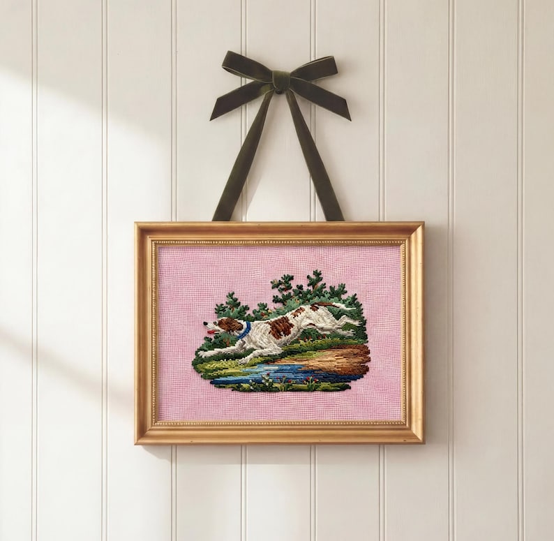 May include: Framed needlepoint art featuring a running dog with brown spots, a blue collar, and a pink background. The artwork is set in a gold frame and hangs from a dark green velvet ribbon. The dog is running near a body of water and trees.