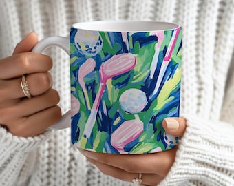 May include: White ceramic mug featuring a colorful golf-themed design. The pattern includes golf clubs, golf balls, and abstract strokes of pink, blue, green, and white. The mug is held by a person wearing a white sweater.