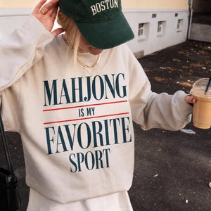 Puede incluir: Una sudadera de cuello redondo color crema con el texto azul oscuro "MAHJONG IS MY FAVORITE SPORT" y finas líneas horizontales rojas encima y debajo de la palabra "IS MY". La sudadera se combina con una gorra de béisbol verde oscuro con "BOSTON" en blanco.