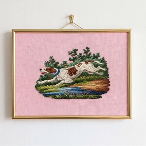 May include: A framed needlepoint artwork featuring a running dog with brown and white spots, wearing a blue collar. The dog is set against a backdrop of green trees, a blue pond, and a pink background. The frame is gold.