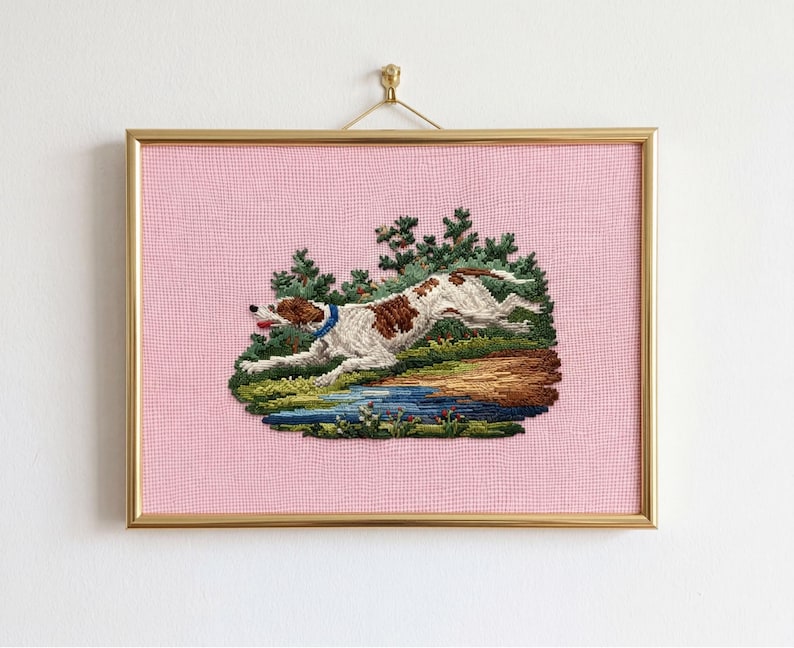 May include: A framed needlepoint artwork featuring a running dog with brown and white spots, wearing a blue collar. The dog is set against a backdrop of green trees, a blue pond, and a pink background. The frame is gold.