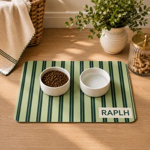May include: A rectangular pet feeding mat featuring a light green background with dark green vertical stripes. The mat includes two white ceramic bowls, one filled with brown kibble and one with water. The text RAPLH is printed in dark green.