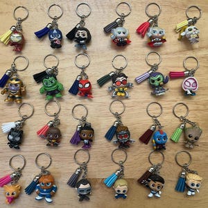 Peut inclure: Une collection de porte-clés de personnages avec des anneaux métalliques et des pompons colorés. Les porte-clés présentent diverses figurines de super-héros et de dessins animés aux couleurs vives, dont un chat, un Hulk vert et un personnage en costume bleu.