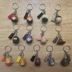 May include: A collection of thirteen metal keychains, each featuring a small character figurine and a colorful fabric tassel. The characters include various figures with distinct features, colors, and expressions, all attached to silver-toned rings.