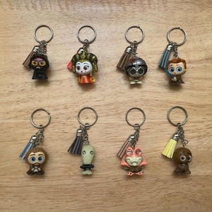 May include: Eight character keychains with silver rings and decorative tassels. The keychains feature various colorful character figurines. The figurines include a black figure, a figure with a crown, and a figure with a helmet.