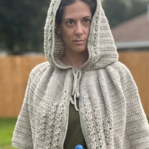 以下が含まれることがあります： グレーの編み物で作られたフード付きケープ。編み込みのデザインが施されています。首元には紐で絞るデザインです。