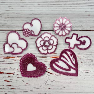 May include: A collection of decorative embellishments in various shapes, including hearts, flowers, and a cross. Each piece features a white interior with a glittery pink border. The embellishments are arranged on a light-colored wooden surface.