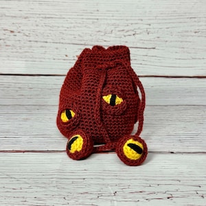 Pode incluir: Uma bolsa de dados de crochê vermelha com olhos amarelos e pretos. A bolsa tem um fecho de cordão e é decorada com três olhos amarelos e pretos.