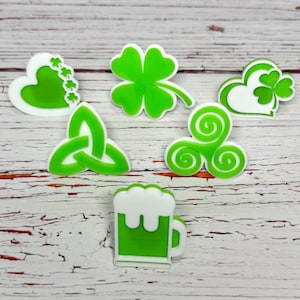 May include: A collection of green and white St. Patrick's Day themed charms. The charms include a shamrock, a beer mug, a Celtic knot, and other Irish symbols. The charms are made of a glossy material and are set against a light wood grain background.