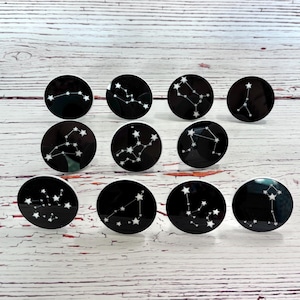 May include: A collection of ten black and white round cabinet knobs. Each knob features a different constellation design in white, set against a black background. The knobs are arranged on a light wood surface.