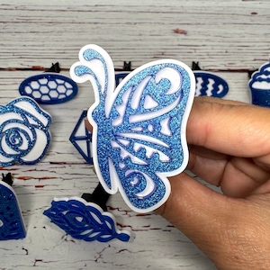 May include: A close-up of several decorative blue and white embellishments. The primary focus is a glittery blue butterfly with intricate cut-out details. Other shapes include a rose, a diamond, and an oval with a honeycomb pattern.