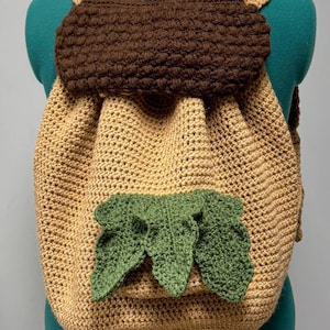 Może przedstawiać: Plecak w kształcie żołędzia wykonany na szydełku z brązowej, beżowej i zielonej przędzy. Plecak ma duży kształt żołędzia z brązową górą i beżowym korpusem. Na dole plecaka znajdują się trzy zielone liście.