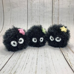 May include: Three handmade, black, fuzzy plush creatures with large white eyes and small, colorful star-shaped accents. The plush toys are arranged in a row against a light wood-grain background. The star accents are pink, green, and yellow.