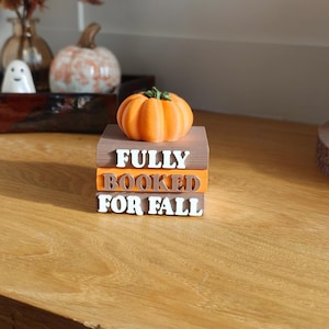 Può includere: Una pila decorativa di tre blocchi rettangolari che ricordano dei libri, colorati in tonalità marrone e arancione. Il testo bianco sui blocchi recita FULLY BOOKED FOR FALL. Una piccola zucca arancione brillante con un picciolo verde è posta in cima.