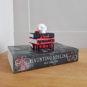 May include: A stack of three miniature black and white books topped with a white skull and a red rose. The books feature the red text Dark Romance. This stack rests on a large black book titled Haunting Adeline by H.D. Carlton with red accents.