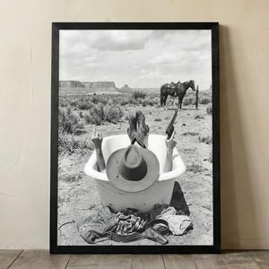 Pode incluir: Uma impressão emoldurada a preto e branco que retrata uma pessoa numa banheira numa paisagem desértica. A pessoa usa um chapéu de cowboy, segura um revólver e faz um gesto com a mão. Um cavalo está ao fundo perto de um poste de madeira.