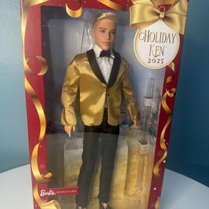 May include: Holiday Ken 2025 doll in a display box. The doll is dressed in a gold tuxedo jacket, black trousers, and a bow tie. The box is red with gold accents and a gold ribbon. The Barbie Signature logo is visible.