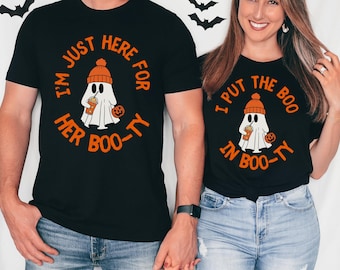 May include: Black Halloween t-shirts with matching designs. One shirt reads "I'M JUST HERE FOR HER BOO-TY" and the other says "I PUT THE BOO IN BOO-TY". Each shirt features a ghost wearing an orange hat and holding a drink and a pumpkin.