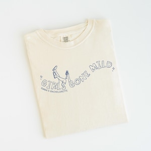 May include: A cream-colored short-sleeve t-shirt featuring a dark blue line art graphic of legs in the air and a hand holding a glass. The text reads GIRLS GONE MILD and EMMA'S BACHELORETTE, accented by small star symbols on each side of the text.