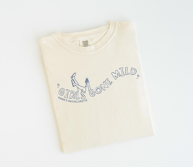 May include: A cream-colored short-sleeve t-shirt featuring a dark blue line art graphic of legs in the air and a hand holding a glass. The text reads GIRLS GONE MILD and EMMA'S BACHELORETTE, accented by small star symbols on each side of the text.