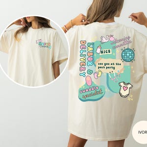 May include: Ivory-colored t-shirt with colorful graphics on the back, including text that reads "Labor & Delivery," "NICU," and "Swaddle Specialist." The front features the text "Nurse Emma."