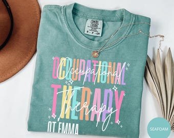 May include: A seafoam green short-sleeve t-shirt featuring the text Occupational Therapy in colorful block letters, with the name OT EMMA printed below. The shirt includes a white script overlay and small star graphics, with a Comfort Colors label.