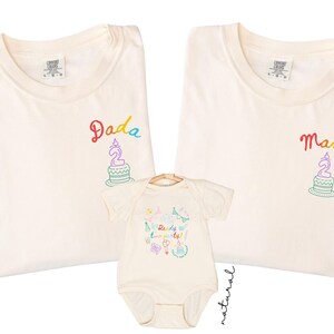 May include: Two ivory t-shirts and one natural baby bodysuit. The t-shirts feature embroidered text reading Dada and Mama above a birthday cake graphic. The bodysuit displays colorful party graphics and the text Ready two party!