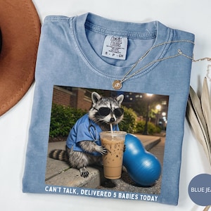 May include: A light blue Comfort Colors t-shirt featuring a graphic of a raccoon wearing sunglasses and a blue medical coat, drinking iced coffee. The text "CAN'T TALK, DELIVERED 5 BABIES TODAY" is printed at the bottom.