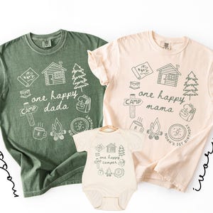 May include: Three camping-themed shirts in moss green, ivory, and ivory. The shirts feature camping-related illustrations and the phrases "one happy dada", "one happy mama", and "one happy camper". The shirts are for a family.