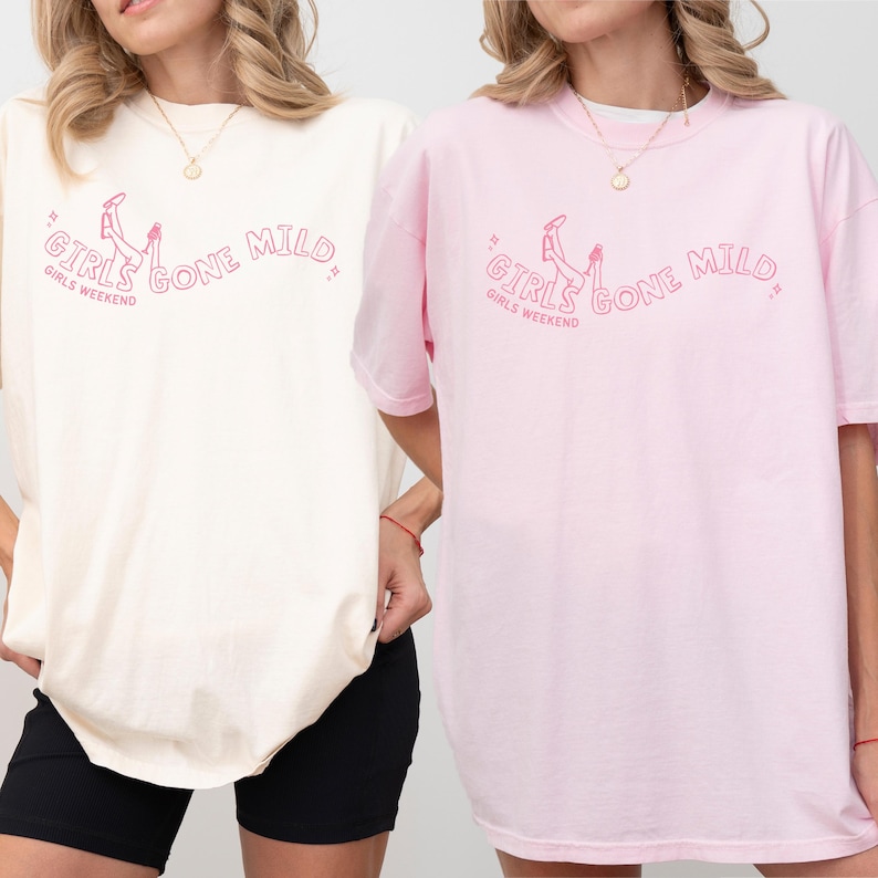 May include: Two short-sleeved crew neck t-shirts, one in cream and one in light pink, each featuring a pink graphic print of legs and the text GIRLS GONE MILD GIRLS WEEKEND. The shirts have a relaxed fit and are made of a solid-colored cotton fabric.