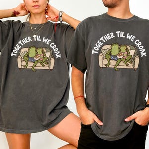 May include: A charcoal grey t-shirt featuring a graphic of two green frogs sitting on a couch eating from a bowl. The text "TOGETHER TIL WE CROAK" is printed in white above the illustration. The shirt has a relaxed fit and a vintage-washed appearance.