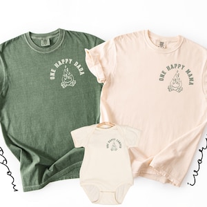May include: Three matching t-shirts and a onesie. The shirts are moss green and ivory, and the onesie is ivory. Each item has a graphic of a campfire with the text "ONE HAPPY DADA", "ONE HAPPY MAMA", and "ONE HAPPY CAMPER".