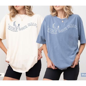 May include: Two oversized t-shirts in ivory and blue jean colors, each featuring a graphic print of legs in the air with the text "GIRLS GONE MILD" and "EMMA'S BACHELORETTE" in a curved layout, paired with black shorts.