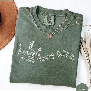 May include: A moss green short-sleeve t-shirt with a white graphic print that reads GIRLS GONE MILD and GIRLS WEEKEND. The design includes a line drawing of legs and a glass. The shirt features a Comfort Colors brand label on the inner collar.
