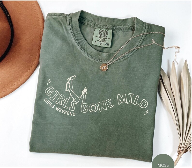 May include: A moss green short-sleeve t-shirt with a white graphic print that reads GIRLS GONE MILD and GIRLS WEEKEND. The design includes a line drawing of legs and a glass. The shirt features a Comfort Colors brand label on the inner collar.