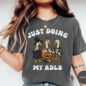 May include: Dark gray t-shirt with the text "JUST DOING MY ADLS" in white. The graphic features three cartoon ducks dressed in Halloween costumes: a witch, a pumpkin, and a skeleton. The shirt is a comfortable, casual style.