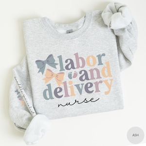 May include: A light grey sweatshirt with the text "labor and delivery nurse" in colorful letters with a bow and baby footprints.