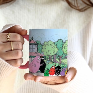 Könnte beinhalten: Eine rosa-weiße Keramiktasse mit einer Cartoon-Illustration einer Gruppe von Figuren in einer Parklandschaft. Die Figuren sind bunt und haben unterschiedliche Formen und Größen. Die Tasse hat einen schwarzen Henkel.