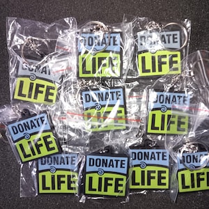 May include: A selection of keyrings, individually packaged in clear plastic. Each keyring displays the words "DONATE LIFE" in a striking blue and neon green design, complete with a silver key ring.