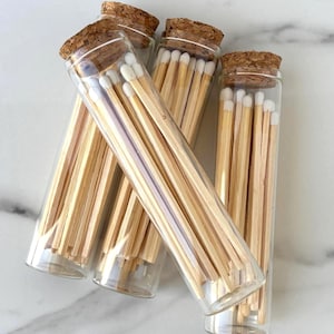 May include: Four clear glass tubes filled with wooden matches. Each tube has a cork stopper. The match heads are white. The wooden matchsticks are a light brown color. The tubes are arranged at a slight angle.