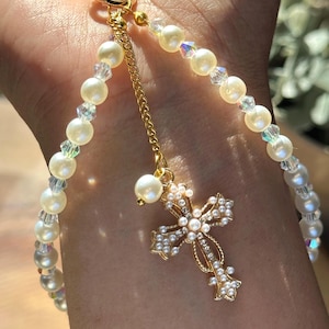 May include: A gold-colored keychain with a pearl and crystal beaded strap. A gold cross pendant with pearl and crystal accents hangs from a gold chain. The cross is adorned with small pearls and crystals.