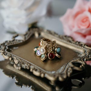 May include: A gold ring with multiple colourful gemstone charms sits on a decorative, antique-style tray. The ring has a textured design and is adorned with various coloured stones, including red, blue, and pearl-like accents. Soft focus flowers are in the background.