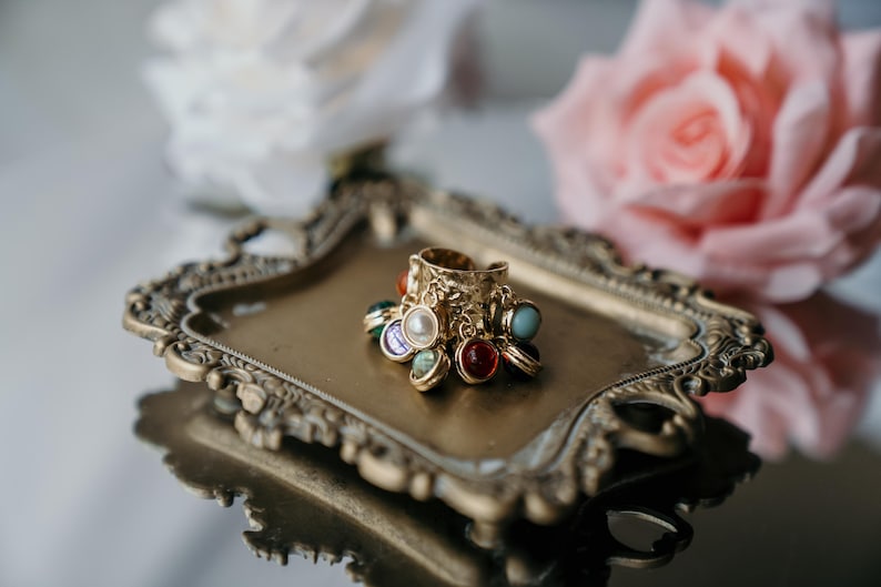May include: A gold ring with multiple colourful gemstone charms sits on a decorative, antique-style tray. The ring has a textured design and is adorned with various coloured stones, including red, blue, and pearl-like accents. Soft focus flowers are in the background.