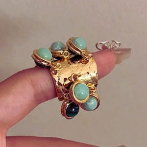 May include: A gold-coloured statement ring with a textured band and multiple dangling charms. The charms feature round, light green and dark green stones. The ring is a unique piece of handmade jewellery.