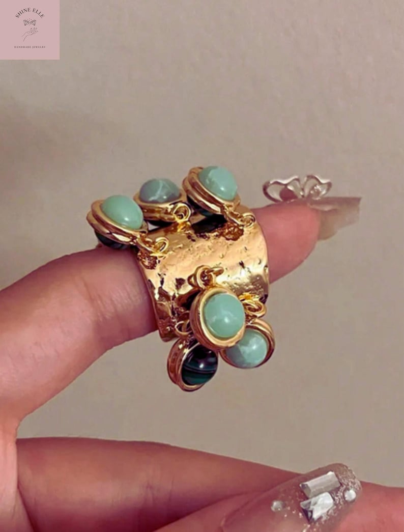 May include: A gold-coloured statement ring with a textured band and multiple dangling charms. The charms feature round, light green and dark green stones. The ring is a unique piece of handmade jewellery.